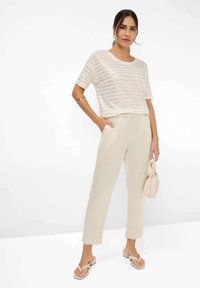 Lichte gestreepte top in beige en wit, gecombineerd met crèmekleurige taps toelopende broeken. Accessoires omvatten een kleine beige handtas en neutrale sandalen.