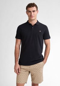 Polo shirt nera a maniche corte con colletto, caratterizzata da un piccolo logo. Abbinata a pantaloni corti beige, decorati da un motivo sottile.