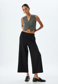 Grijze cropped vest met een V-hals en knoopsluiting, gecombineerd met zwarte wijde broek. Zwarte instapschoenen maken de outfit af.