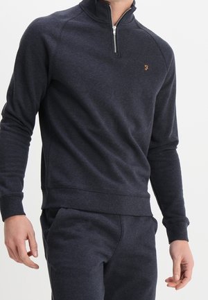 Homme portant un sweat-shirt à fermeture éclair quart de zip bleu marine foncé et un pantalon de jogging assorti avec un petit logo jaune brodé sur la poitrine.