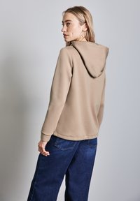 Sudadera con capucha beige hecha de material suave, con un bolsillo canguro y puños acanalados, combinada con pantalones de mezclilla anchos de color oscuro.
