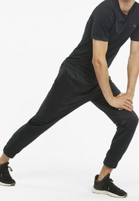 Person in schwarzer Sportkleidung, der das Bein mit den Händen auf dem gebeugten Knie gegen einen schlichten weißen Hintergrund dehnt.