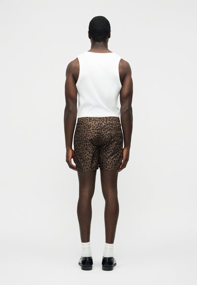 Homme portant un débardeur blanc, un short imprimé léopard, des chaussettes blanches et des chaussures noires, debout de dos sur un fond uni.