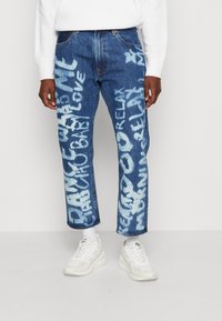 Jeans i denim i mörkblått med ljusblå textdesign. Har rakt snitt och standard fem-ficksstil. Avslutas med vita sneakers.