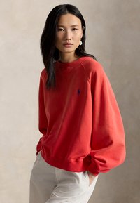 Γυναίκα με μακριά μαύρα μαλλιά φορεί κόκκινο φούτερ με μπλε λογότυπο και άσπρο παντελόνι, στέκεται μπροστά σε απλό μπεζ φόντο.