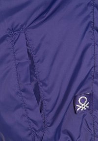 Fialová nylonová tkanina s lesklou texturou, se šitou kapsou a malou vyšitou značkovou cedulkou ve stříbrné barvě v pravém dolním rohu.