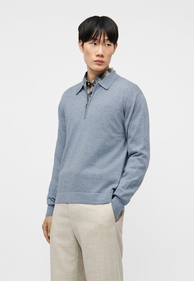 Άνδρας με ανοιχτό μπλε πουλόβερ με φερμουάρ πάνω από πουκάμισο καρό και μπέζ παντελόνι, στέκεται με το ένα χέρι στην τσέπη, κοιτάζοντας προς το πλάι.