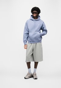 Ανοιχτό μπλε φούτερ με μπροστινή τσέπη, γκρι σορτς και μπεζ αθλητικά παπούτσια. Το μοντέλο φοράει γυαλιά και ανοιχτό μπλε κάλτσες, στέκεται μπροστά σε λευκό φόντο.