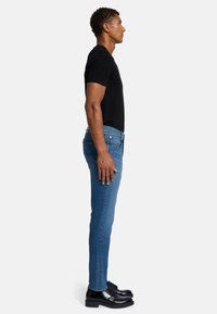 Jeans in denim blu con vestibilità slim, abbinati a una t-shirt nera. Il modello indossa scarpe eleganti nere, mostrando un look casual ma curato.