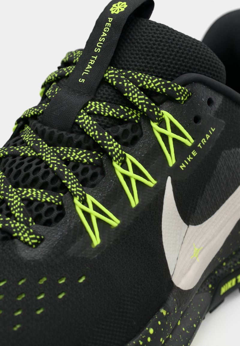 Zapato atlético negro con parte superior de malla texturizada, detalles reflectantes plateados, cordones verdes vibrantes y suela moteada. Diseñado para correr en senderos.