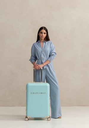 Light blue hard-shell suitcase with a glossy finish, silver hardware, and four wheels. Model wearing matching oversized outfit stands beside it.