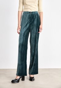 Pantalons larges plissés en teal dans un tissu doux et texturé, avec une taille ajustée et un bouton décoratif en or à l'avant.