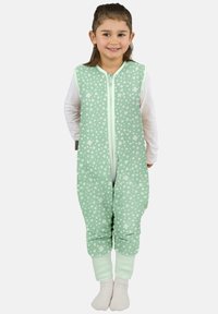 Lächelndes junges Mädchen, das einen ärmellosen Jumpsuit mit mintgrünem Blumenmuster über einem weißen Langarmshirt und weißen Socken trägt, vor einem einfarbigen Hintergrund steht.