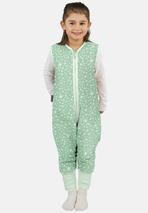 Lächelndes junges Mädchen, das einen ärmellosen Jumpsuit mit mintgrünem Blumenmuster über einem weißen Langarmshirt und weißen Socken trägt, vor einem einfarbigen Hintergrund steht.