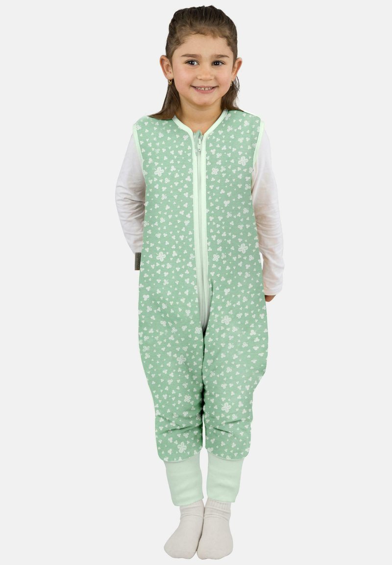 Lächelndes junges Mädchen, das einen ärmellosen Jumpsuit mit mintgrünem Blumenmuster über einem weißen Langarmshirt und weißen Socken trägt, vor einem einfarbigen Hintergrund steht.