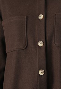 Giacca di lana marrone scuro con tre bottoni chiari e due grandi tasche sul petto, mostrando i dettagli della chiusura anteriore con bottoni.