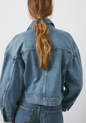 Personne aux longs cheveux roux attachés en queue de cheval, portant une veste en denim bleue et un jean assorti, vue de dos sur un fond blanc.