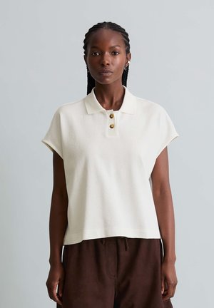 Femme noire aux cheveux tressés portant un polo blanc sans manches et un pantalon marron, debout devant un fond gris clair uni.