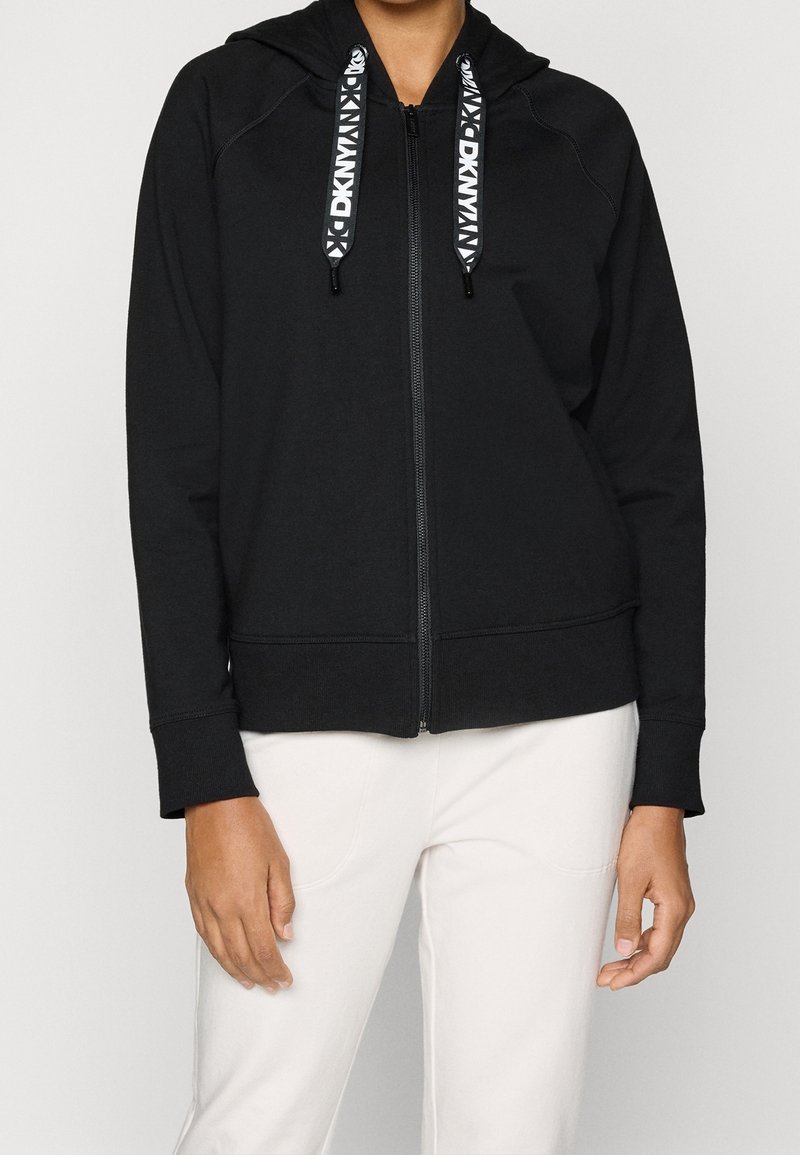 Sweat à capuche noir zippé avec cordons de marque DKNY, porté avec un pantalon blanc sur une personne visible du cou aux hanches.