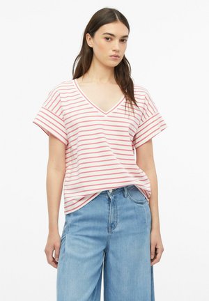Jeune femme portant un haut court à rayures blanches et roses avec un col en V et un jean ample bleu clair, debout devant un fond uni.