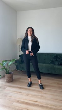 Casaco curto preto sobre uma blusa branca, combinado com calças pretas justas e mocassins pretos. Sofá texturizado verde e planta ao fundo.