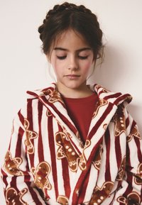 Accappatoio a righe rosse e bianche con motivi di omini di pan di zenzero, tessuto morbido e peloso, con cappuccio, indossato sopra una maglietta rossa.