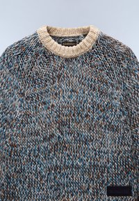 Sweter dzianinowy z okrągłym, beżowym kołnierzem. Włóczka w kolorach brązowym, niebieskim i białym tworzy teksturowany wzór. Szczegół z czarną metką.
