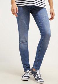 Jeans skinnys de mezclilla azul con un lavado desvanecido, con costuras curvadas a lo largo de los muslos, combinados con zapatillas negras y blancas.