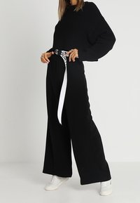 Maglione cropped nero con texture a costine, abbinato a pantaloni a gamba larga neri con una cintura bianca e scritte nere, completato con scarpe da ginnastica bianche.