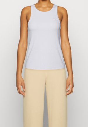 Femme portant un débardeur gris clair sans manches et un pantalon beige à jambes larges, debout devant un fond clair uni.