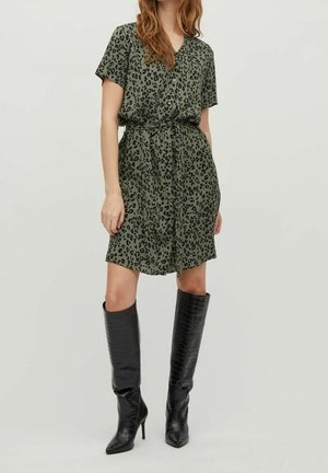 Femme portant une robe verte à manches courtes avec ceinture à imprimé léopard et des bottes noires à talons montant jusqu'aux genoux, debout devant un fond uni.