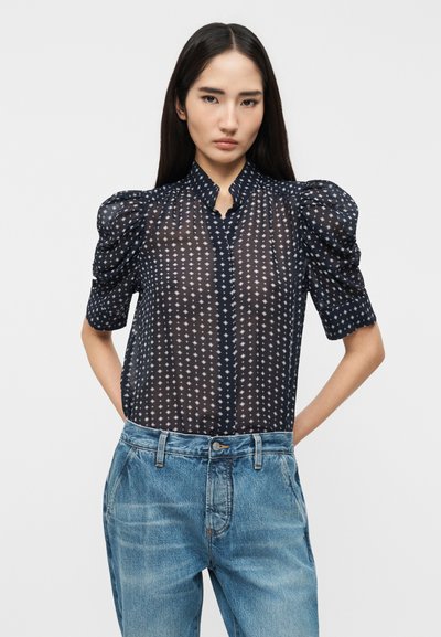 Γυναίκα που φοράει διαφανή μπλε μπλούζα με φουσκωτά μανίκια και λευκά γεωμετρικά σχέδια, συνδυασμένη με ανοιχτό μπλε τζιν παντελόνι με ψηλή μέση.