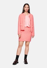 Rosa strukturierte Jacke mit passendem Rock, über einer blassrosa Bluse geschichtet. Das Model trägt schwarze Socken und dunkelbraune Schuhe mit Quasten-Details.