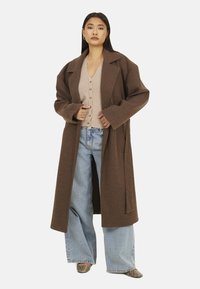Cappotto in lana marrone con rever ampi; abbinato a un cardigan beige con bottoni e jeans a gamba larga azzurri chiari. Tessuto strutturato e vestibilità ampia.