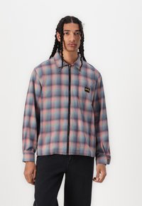 Men's zip-up plaid jacket in blue and pink tones, featuring a collared neckline, long sleeves, and left chest logo patch. Woven fabric texture.