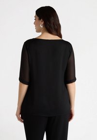 Top nero con maniche trasparenti a tre quarti, scollatura rotonda e vestibilità rilassata. Il tessuto ha una texture liscia. Vista da dietro.