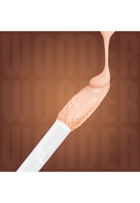 Fond de teint liquide beige coulant sur une éponge applicatrice de maquillage douce à manche blanc sur un fond marron.