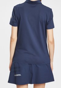 Marinblå piké med korta ärmar, slät tygkvalitet och klassisk krage. Matcherad med en marinblå kjol som har en logotyp och diskreta ränder.