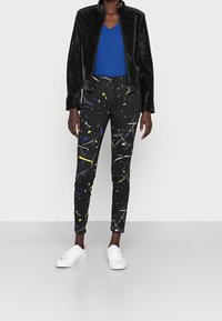 Jeans negros con un patrón de salpicaduras blancas, amarillas y azules, combinados con una camiseta azul de cuello en V y una chaqueta negra texturizada, completado con zapatillas blancas.