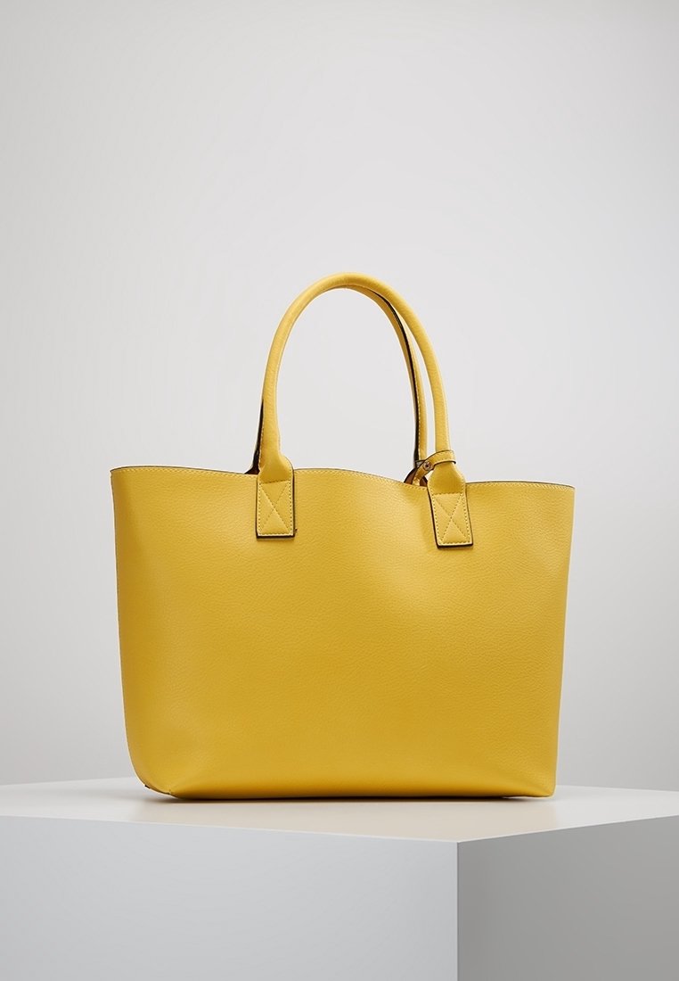 Sac cabas en cuir jaune avec doubles poignées placé sur un piédestal blanc sur un fond uni gris clair.