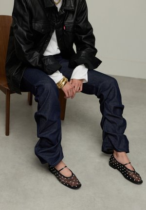 Zwarte leren jas, witte shirt, donkere spijkerbroek en zwarte mesh flats met witte stippen. Model zittend op een houten stoel met een neutrale achtergrond.