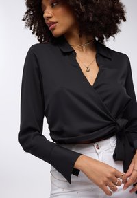 Blusa nera avvolgente con maniche lunghe e dettaglio di cravatta in vita. Tessuto liscio, colletto a risvolto e polsini svasati. Abbinata a jeans bianchi.