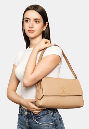 Giovane donna con capelli castani lisci tiene una borsa in pelle beige sulla spalla, indossa una maglietta bianca e jeans blu su sfondo neutro.