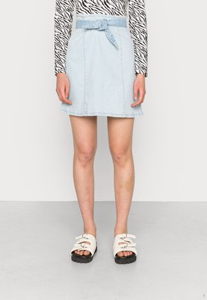Saia de ganga azul clara com cinto de laço, design em A, com bainha crua, apresentando costuras frontais. Combinada com sandálias brancas de tiras e solas pretas.