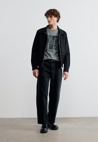 Νέος άντρας με σγουρά μαλλιά, φορώντας μαύρο μπουφάν, γκρι μπλούζα με σχέδιο, μαύρο παντελόνι και μαύρα παπούτσια, στέκεται μπροστά σε λευκό, απλό τοίχο.