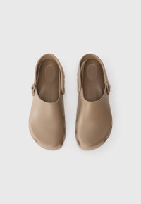 Et par beige slip-on klipklapper med rygsrok, rund tå og polstrede såler på en ensfarvet hvid baggrund.
