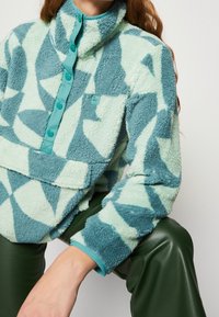 Suéter de forro polar en menta y verde azulado, con un patrón abstracto, cuello alto, cierre de botones y un bolsillo parche. Textura suave y ajuste relajado.