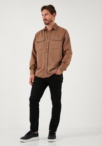 Smeđa košulja s dugim rukavima i dvjema džepovima na prsima, crne slim-fit hlače i crne tenisice s bijelim đonom, stoji naslonjena na praznu pozadinu.