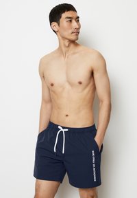 Marinblå simbyxor med vit dragsko, med sidofickor och vertikal vit text "MARC O'POLO EST. I STOCKHOLM" på vänster ben.