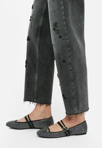 Zwart-witte ballerina's met strassdetails, twee zwarte banden over de voetboog, gedragen met versleten grijze jeans met versieringen.